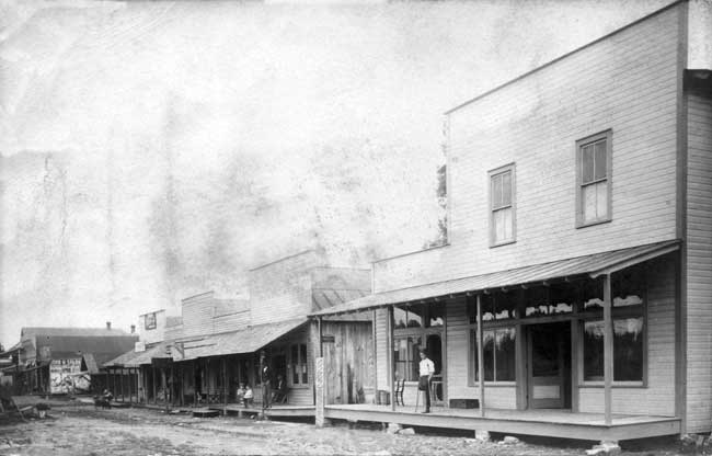 Dirt road through town with wood framed buildings and covered porches, man with one leg on crutches
