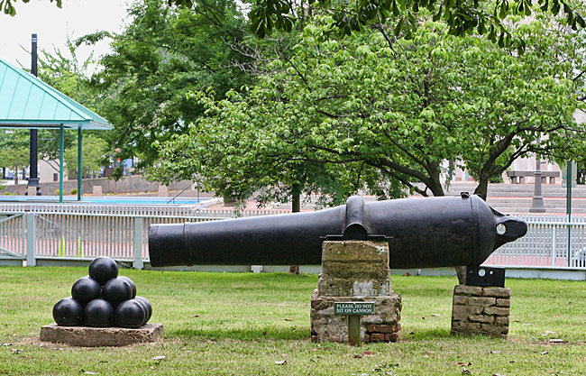 Side view of cannon balls and cannon on brick pedestals inside fence on grass with trees and buildings in the background