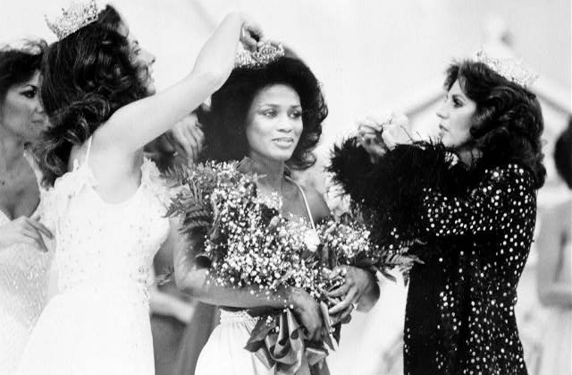 Black woman in white dress with bouquets being crowned by two white women also crowned
