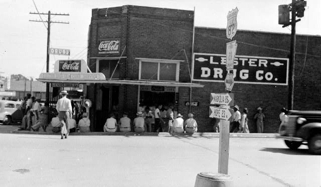 Men gathered seated on curb and standing outside multistory brick building with sign saying "Lester Drug Company" and "coca-cola"