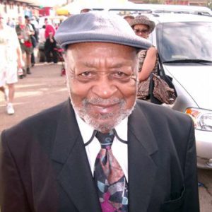 older black man in suit and abstract-pattern tie and cap smiling near pedestrians cars and buildings