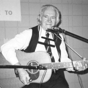 Older white man in vest tie smiling poses with guitar and harmonica  two microphones