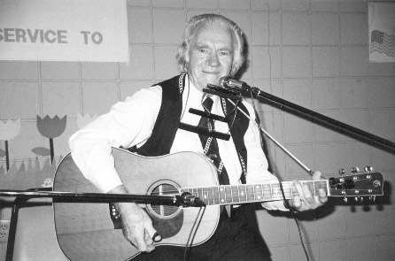 Older white man in vest tie smiling poses with guitar and harmonica  two microphones