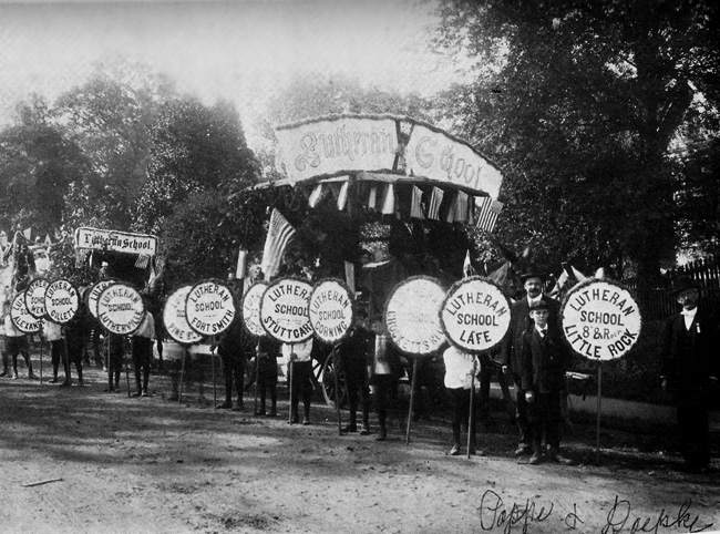 Lutheran School parade along wooded road with participants holding round signs representing various school cities, photograph hand-signed "Pappe and Doepke"