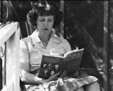 White woman seated reading on porch in blouse and skirt