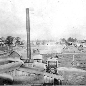 Wooden mill buildings with a tall smokestack and rail yard in the background