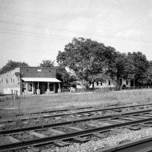 Brick building with front awning trees and neighboring wood frame home seen from railroad tracks