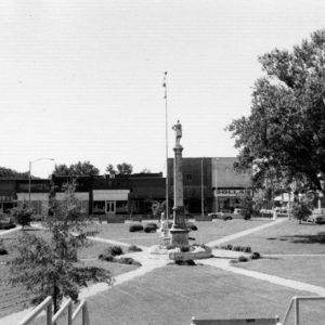 Park with flagpole and central monument figure on column with three intersecting sidewalks with trees and surrounding businesses