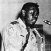 African American man in state trooper uniform speaks into several microphones