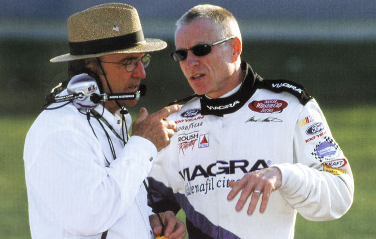 White man in a straw hat talking to a white man in a "Viagra" fire suit