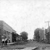 Dirt road lined with brick buildings, one with sign reading "Farmers Bank," wooden plank sidewalks, pedestrians