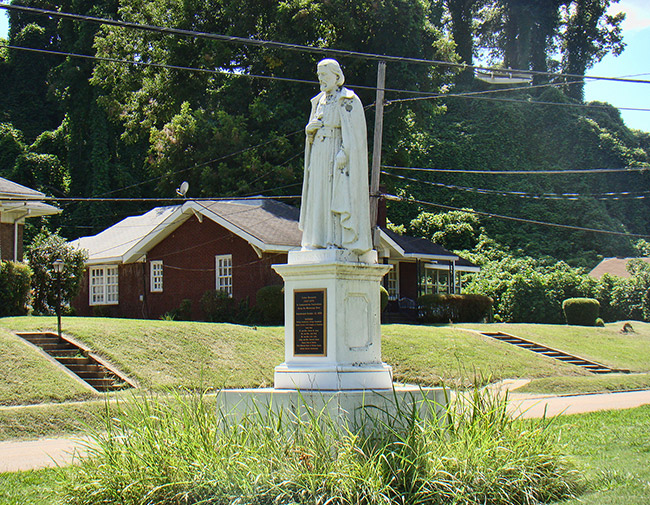 Statue of man in robes standing on a pedestal with plaque on street corner in residential neighborhood