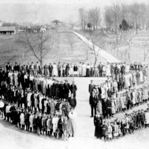 Three slanted rows of people lined up by age with center aisle and a back fourth row and distant town