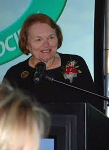 White woman with corsage speaking at a lectern