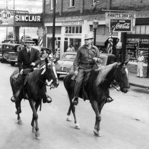 Two white men in hats on horseback on street passing observers outside gas station barber shop and grocery