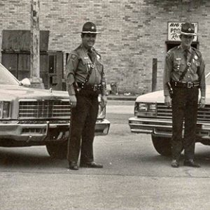 Two white policemen in uniform with patrol cars in parking lot