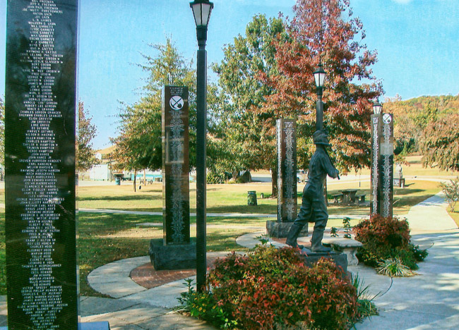 Statue of man with pick ax surrounded by engraved columns on walking path in park with lamps and trees in the background