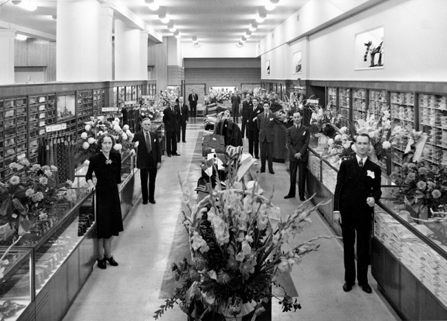 Dressed up department store workers pose in aisles by glass cases racks displays flower bouquets