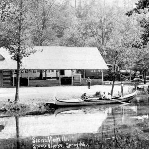 Riverside cabin in woods with people, canoe, and carriage, hand-labeled "Scene Monte Ne."