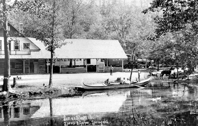 Riverside cabin in woods with people, canoe, and carriage, hand-labeled "Scene Monte Ne."
