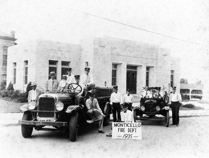 Group portrait of firemen with fire truck and vehicle with sign "Monticello Fire Dept 1935" outside brick building