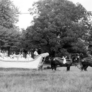 Group of white women in dresses and men in top hats being pulled in a giant sleigh parade float by horses