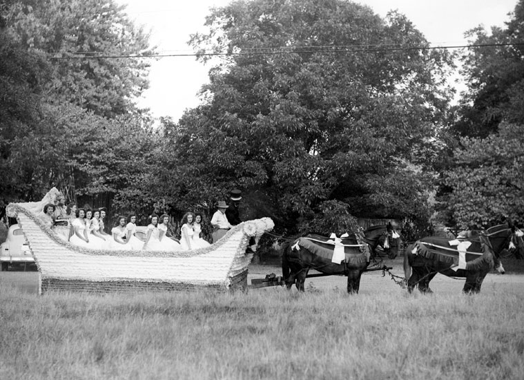 Group of white women in dresses and men in top hats being pulled in a giant sleigh parade float by horses