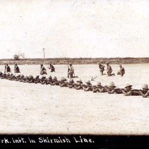 Long row prone soldiers in dirt field aiming rifles kneeling officers behind caption "skirmish line"