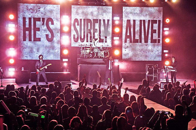 Musicians performing on stage to crowd with "He's Surely Alive" background behind them