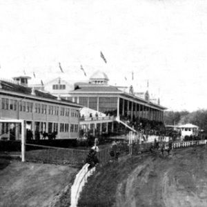 horses and jockeys on track and large buildings with flags and spectators behind