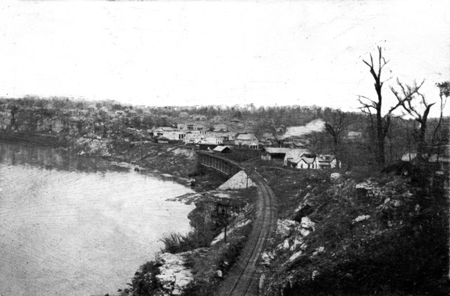 Riverside town view from wooded ridge along railroad tracks with bridge and curving shoreline