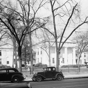 White building with four columns and two wings behind an iron fence with trees and parked cars on Markham Street