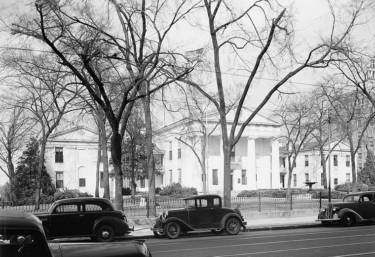 White building with four columns and two wings behind an iron fence with trees and parked cars on Markham Street