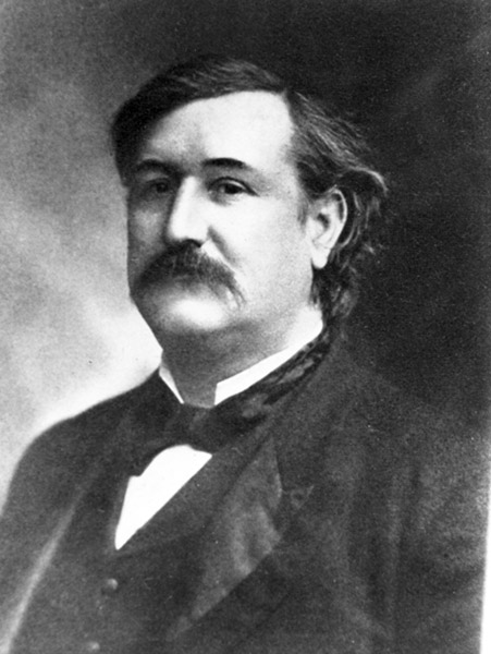 Portrait photo of a white man with a dark mustache brushed hair solemn expression in suit and bow tie