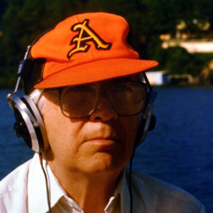 white man by lake squinting under crooked orange baseball cap with an A on it and large glasses wearing headphones