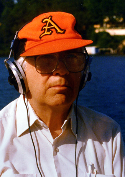white man by lake squinting under crooked orange baseball cap with an A on it and large glasses wearing headphones
