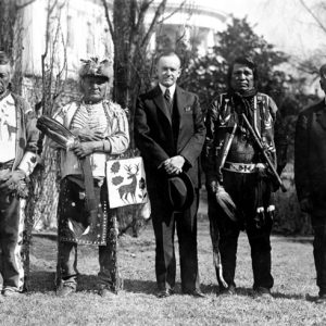 White man in suit holding his hat standing outside multistory house with two Native American men in traditional clothing on each side of him