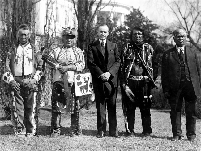 White man in suit holding his hat standing outside multistory house with two Native American men in traditional clothing on each side of him
