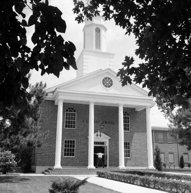 man standing with his arms outstretched on covered porch of multistory brick building with steeple and four columns