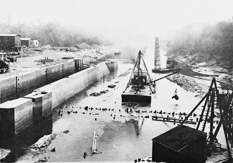 Dam construction in river with flat barge cranes, concrete lock and buildings in background