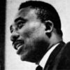 profile view of black man speaking with short hair and mustache wearing a suit and tie