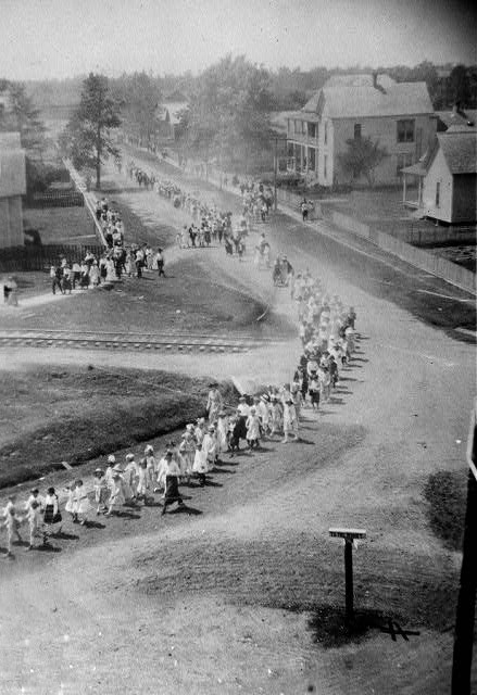 Aerial view of a parade led by school children passing through town by homes over railroad tracks