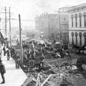 Busy street with men on sidewalk, horse-drawn wagons, road construction, brick buildings