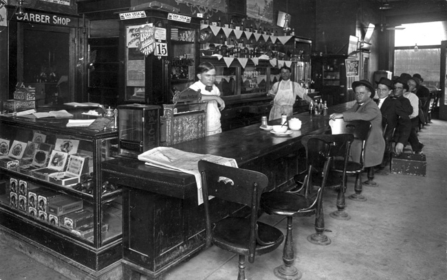 Cafe interior with two waiters, men at bar, cigar case, side doorway marked "Barber Shop"