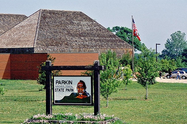 Wooden sign with Native American man on it with a brick building with a wooden roof behind it and flag pole