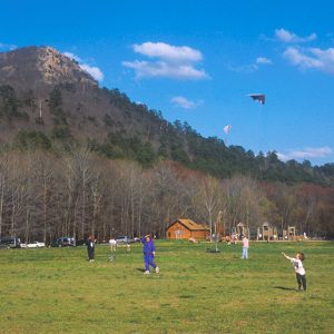 Men women and children playing in field with cabin building and at base of tree covered mountain