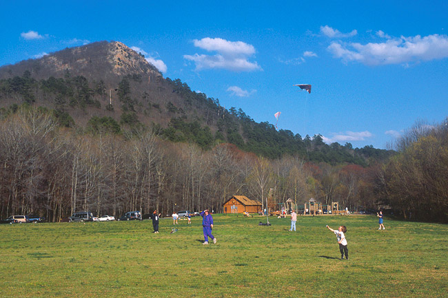 Men women and children playing in field with cabin building and at base of tree covered mountain