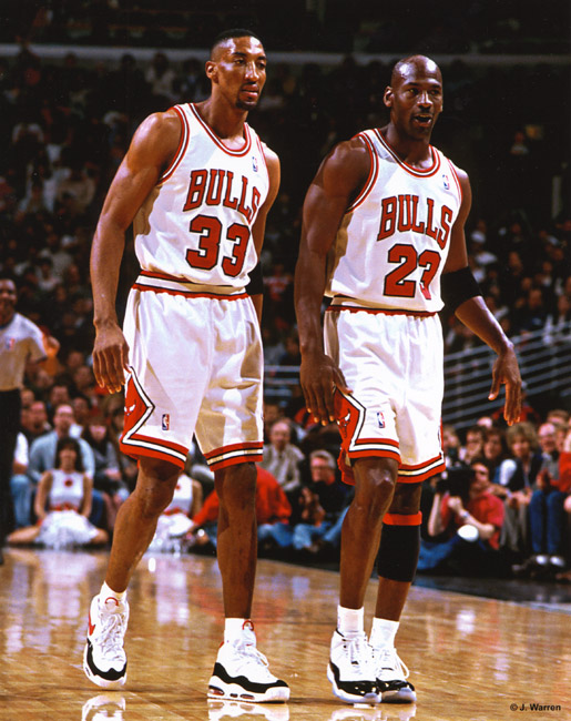 Two black men walking together on the court in NBA Bulls uniforms numbers 33 and 23 with fans in background