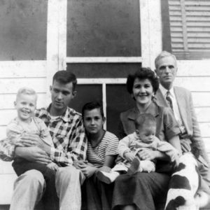White family with 2 men, 1 woman, 3 children, and dog outside wood frame house
