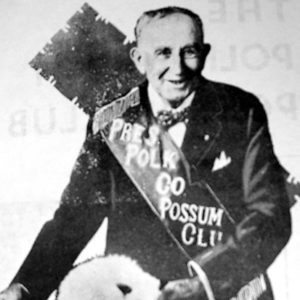 white man in sash "Pres. Polk Co. Possum Club" poses with an opossum